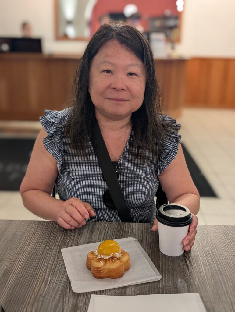Ellen smiling at a cafe table with a coffee and a flower-shaped Le Carousel pastry topped with a mango-pineapple dome and coconut.