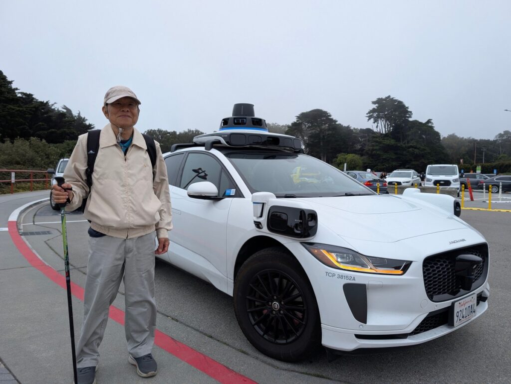 Allan, 82, standing beside a white Waymo driverless Jaguar taxi with roof-mounted sensors in a foggy Golden Gate parking lot.