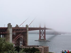 The Golden Gate Bridge with its south tower vanishing into thick fog, Fort Point below and walkers on the path.