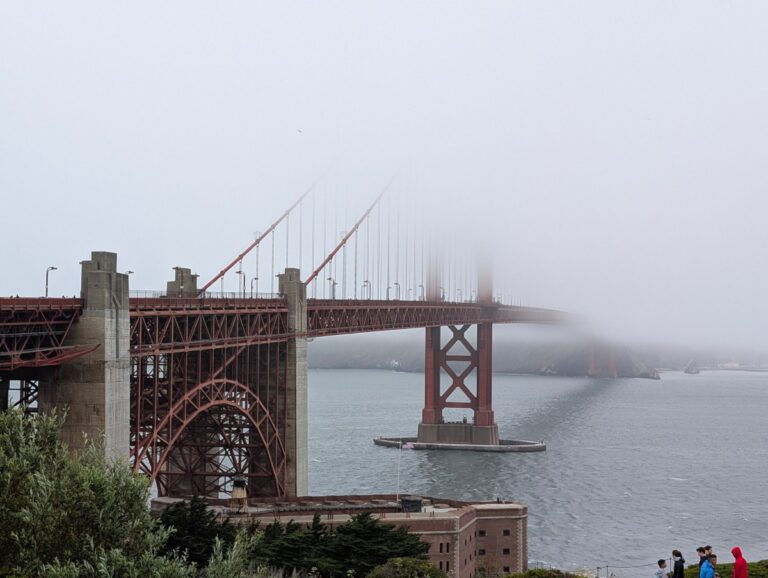 The Golden Gate Bridge with its south tower vanishing into thick fog, Fort Point below and walkers on the path.