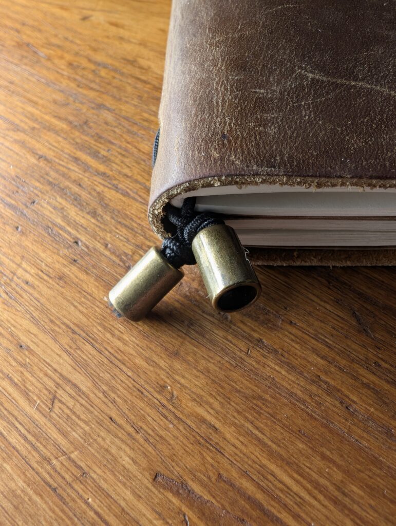 Close-up of the brass bobble cord ends and black elastic closure on the Wanderings A6 traveller's notebook.