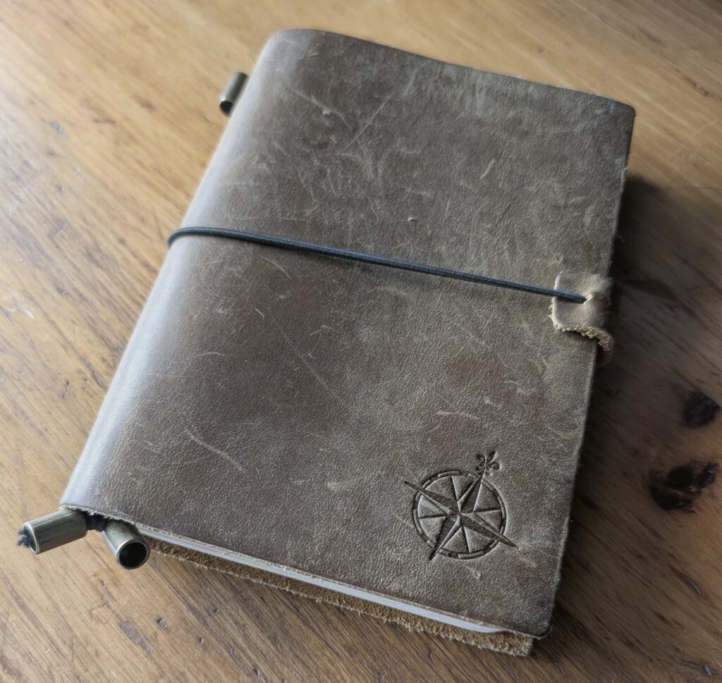 The Wanderings A6 traveller's notebook, closed, showing its distressed brown leather cover embossed with a compass rose and held shut by a black elastic band.